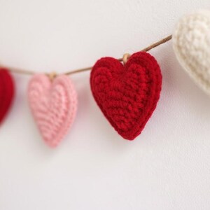 Peut inclure: Une guirlande de c&oelig;urs crochet&eacute;s rouges, roses et blancs est suspendue sur un fond blanc. Les c&oelig;urs sont enfil&eacute;s sur une ficelle naturelle, cr&eacute;ant une esth&eacute;tique charmante et artisanale. Parfait pour la Saint-Valentin.