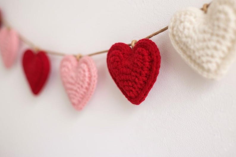Peut inclure: Une guirlande de c&oelig;urs crochet&eacute;s rouges, roses et blancs est suspendue sur un fond blanc. Les c&oelig;urs sont enfil&eacute;s sur une ficelle naturelle, cr&eacute;ant une esth&eacute;tique charmante et artisanale. Parfait pour la Saint-Valentin.