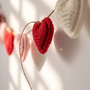 Peut inclure: Une guirlande de c&oelig;urs crochet&eacute;s rouges, roses et blancs est suspendue contre un mur blanc. Les c&oelig;urs sont enfil&eacute;s sur une fine ficelle marron, cr&eacute;ant une pi&egrave;ce d&eacute;corative charmante et faite &agrave; la main. L'&eacute;clairage projette des ombres douces.