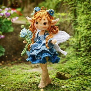 Könnte beinhalten: Eine handgefertigte Häkelfee-Puppe mit orangefarbenem Haar, blauen Blumenakzenten und weißen Flügeln. Die Puppe trägt ein blaues Kleid und hält einen Strauß blauer Blumen. Die Puppe steht auf grünem Moos.