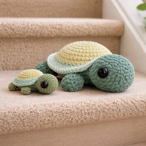 Könnte beinhalten: Zwei gehäkelte Schildkröten-Plüschtiere, eine große und eine kleine, sitzen auf einer beigefarbenen, mit Teppichboden bedeckten Oberfläche. Die Schildkröten haben grüne Körper, gelb-grüne Panzer und große schwarze Augen. Die größere Schildkröte ist etwa 25 cm lang.