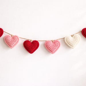 Peut inclure: Une guirlande de six c&oelig;urs crochet&eacute;s, rouges, roses et blancs, suspendus &agrave; une ficelle naturelle. Les c&oelig;urs sont espac&eacute;s de mani&egrave;re uniforme sur un fond blanc, cr&eacute;ant une esth&eacute;tique charmante et artisanale.