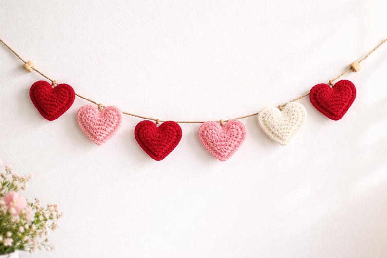 Peut inclure: Une guirlande de six c&oelig;urs crochet&eacute;s, rouges, roses et blancs, suspendus &agrave; une ficelle naturelle. Les c&oelig;urs sont espac&eacute;s de mani&egrave;re uniforme sur un fond blanc, cr&eacute;ant une esth&eacute;tique charmante et artisanale.