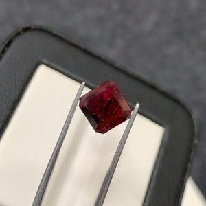 Könnte beinhalten: Ein tiefroter, facettierter Edelstein in quadratischer Form, der von einer silberfarbenen Metallpinzette gehalten wird. Der Stein weist durchscheinende Eigenschaften und scharfe, geometrische Kanten auf, die das Licht auf seiner polierten Oberfläche reflektieren.