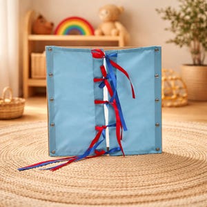 May include: A light blue fabric panel with red and blue ribbons laced through the center. The panel is attached to a wooden frame with silver rivets. The ribbons are tied in bows. The panel is on a woven rug.