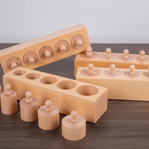 May include: Wooden Montessori cylinder blocks in various sizes. The set includes several rectangular blocks with circular holes and matching cylinders with knobs. The natural wood color and simple design are ideal for early childhood education.
