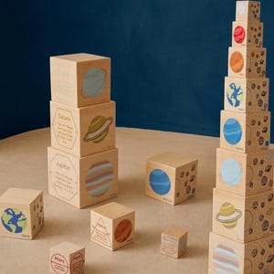 May include: Wooden stacking blocks featuring planets and celestial bodies. Each block is painted with a planet illustration and labeled with its name. The blocks are arranged in a tower and scattered on a neutral surface.