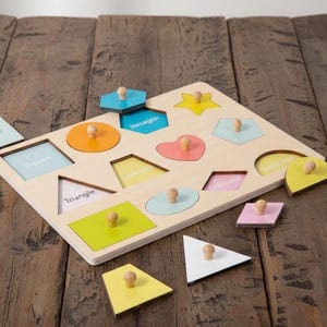 May include: Wooden shape puzzle with various colorful geometric shapes, including a square, triangle, and hexagon. The puzzle pieces have wooden knobs for easy handling. The shapes are in a variety of colors, including blue, yellow, and pink.