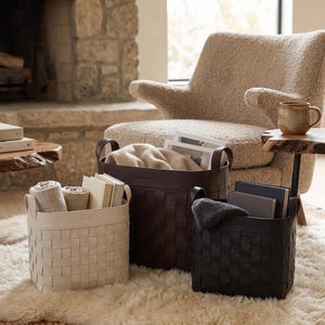 May include: Three square storage baskets with a woven design and side handles, presented in cream, dark brown, and black. The baskets contain various items including folded textiles and books, showcasing a structured, grid-like exterior pattern.