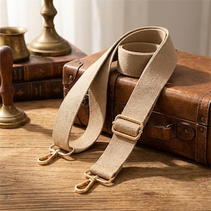 Puede incluir: Una correa de bolso de lona beige con herrajes metálicos dorados. La correa ajustable tiene dos cierres giratorios y una hebilla rectangular. La correa se muestra sobre una superficie de madera con una maleta vintage al fondo.