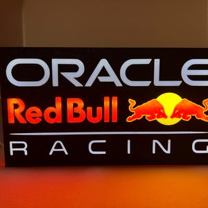 Op de afbeelding: Verlicht bord met het Oracle Red Bull Racing logo. Het bord toont witte letters voor "ORACLE" en "RACING", met "Red Bull" in het rood. Het Red Bull logo, met twee stieren, is ook rood.