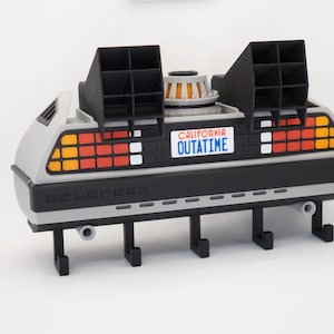 Puede incluir: Un colgador de pared gris y negro con temática DeLorean con una placa que dice "CALIFORNIA OUTATIME". El diseño presenta luces naranjas, rojas y blancas, y ganchos negros. Una pieza divertida y funcional para los fans de la película.