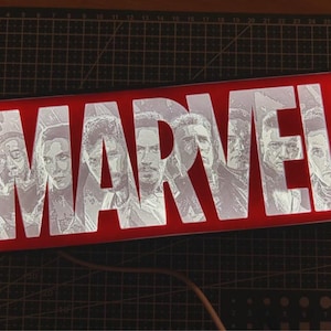 May include: A red rectangular light-up sign with the word "MARVEL" in white, featuring etched portraits of characters. The sign is illuminated, and a power cord is visible.
