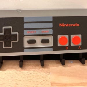 Op de afbeelding: Een grijze en zwarte sleutelhouder ontworpen als een klassieke Nintendo-controller. Het heeft het Nintendo-logo in rood, knoppen en een D-pad. Inclusief vier zwarte haken voor sleutels.
