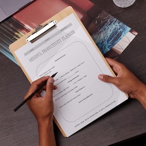 Op de afbeelding: Een houten clipboard met een bedrukt "MINDFUL PRODUCTIVITY PLANNER" vel. Een persoon houdt het clipboard vast en schrijft met een zwarte pen. De planner bevat secties voor intentionele focus, energiebewustzijn en rustige check-ins. De tekst "DOING LESS, WITH INTENTION, IS STILL PRODUCTIVE" staat onderaan.