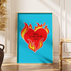 May include: A framed art print featuring a red heart engulfed in flames against a bright blue background. The heart contains the text "BABY, LA VIDA ES UN CICLO" in white. The artwork is displayed in a room with a vase of dried plants.