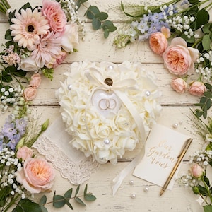 Peut inclure: Un coussin en forme de cœur fait de roses blanches, contenant deux alliances dorées. Le coussin est orné de perles et d'un ruban crème. Des bouquets de roses roses et pêche entourent le coussin, avec un stylo doré.