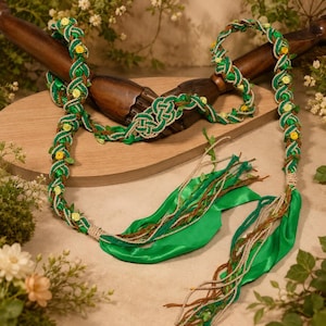 Puede incluir: Un cinturón o guirnalda trenzada con cordones entrelazados en verde y dorado metálico, decorado con pequeñas flores de tela amarilla y hojas verdes. Los extremos terminan en largas cintas de satén verde y finos hilos de color marrón.