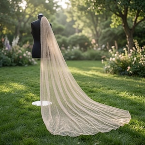 May include: A long, floor-length wedding veil made of sheer, champagne-colored tulle fabric with a subtle shimmering glitter finish. The veil is draped over a black mannequin torso, extending outward to rest on the ground in a soft, flowing train.