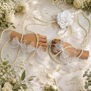 May include: Two wooden mannequin hands are connected by a long, twisted white and gold cord adorned with sheer white organza bows. A large white fabric rose is attached to the cord. A small card with the text Garden Wedding is visible.