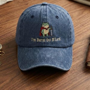 Könnte beinhalten: Blaue Denim-Baseballkappe mit einer Stickerei einer Froschfigur mit Umhang und Schwert. Der Text "I'm Burnt Out M'Lord" ist darunter gestickt. Die Kappe hat einen gebogenen Schirm und einen verstellbaren Riemen.