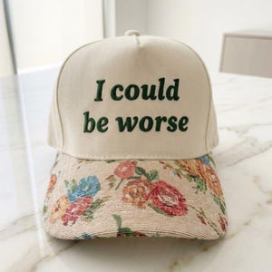 Op de afbeelding: Een beige baseballpet met de geborduurde zin "I could be worse" in groen. De klep heeft een bloemenpatroon met rode, blauwe en gele bloemen.