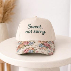 Könnte beinhalten: Cremefarbene Baseballkappe mit dem Text "Sweet, not sorry" in grüner Schrift. Der Schirm hat ein Blumenmuster mit rosa, blauen und gelben Rosen auf cremefarbenem Hintergrund. Die Kappe liegt auf einem weißen Tisch.