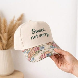 Könnte beinhalten: Cremefarbene Baseballkappe mit dem gestickten Text "Sweet, not sorry". Der Schirm hat ein Blumenmuster mit rosa, blauen und gelben Rosen. Die Kappe wird von einer Hand gehalten.