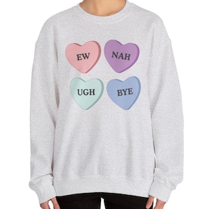 Può includere: Una felpa grigio chiaro con quattro caramelle a forma di cuore rosa, viola, verde menta e blu. Ogni caramella ha una parola: "EW", "NAH", "UGH" e "BYE". La felpa è in stile casual.