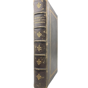 Può includere: Un libro antico intitolato "Bunyan's Pilgrim's Progress" con dorso e copertina in pelle marrone scuro. Il dorso presenta scritte dorate e dettagli decorativi. I bordi del libro mostrano segni di usura, indicando l'età.