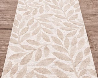 May include: Rectangular beige rug with a repeating leaf pattern. The leaves are a lighter shade of beige, creating a subtle contrast against the off-white background. The rug is placed on a wooden floor.