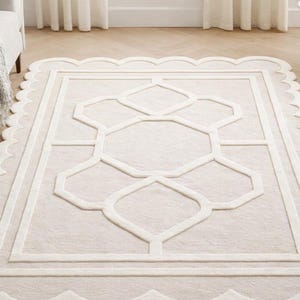 May include: A cream-colored area rug with a scalloped border and a geometric design. The rug features a central rectangular pattern with intersecting lines and shapes, creating a modern aesthetic. The rug is placed on a light-colored wooden floor.