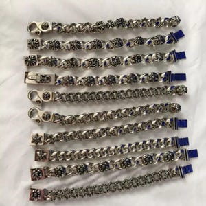 May include: An assortment of silver-toned chain bracelets with various decorative links. Some bracelets feature ornate cross and floral designs, while others have a simple chain link pattern. Each bracelet has a clasp and a blue stone accent.