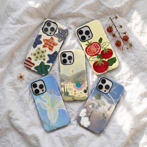 May include: Five smartphone cases featuring diverse designs: one with colorful patterned stars, one with red tomatoes on a lined background, one with a surreal landscape, one with a pale lily on blue, and one with white flowers and a moth.