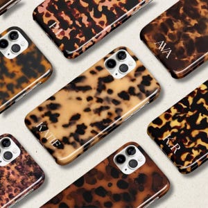 May include: A collection of tortoiseshell-patterned phone cases in various shades of brown and tan. Some cases feature personalized initials in white. The cases have a glossy finish and are designed to fit a smartphone with a triple-lens camera.