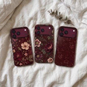 May include: Three rectangular smartphone cases featuring dark burgundy camera modules. The left case displays a floral pattern with light pink and deep red flowers. The center case shows larger blooming flowers. The right case has a subtle tonal floral print.