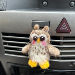 May include: A handmade felt owl figurine with brown, white, and yellow details. The owl has pink cheeks, a yellow beak, and black eyes. The owl is a decorative item.