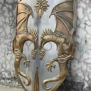 May include: A large, shield-shaped metal piece featuring a polished silver-toned centre and a bronze-toned border. Two symmetrical dragon figures are embossed on the sides, with a star-shaped emblem at the top and a pointed spike at the base.