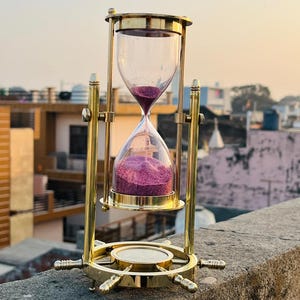 May include: A brass hourglass featuring a ship's wheel base and a compass in the center. The glass bulbs contain vibrant purple sand, with a portion of the sand currently flowing from the top bulb into the bottom bulb through the narrow neck.
