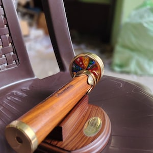 Könnte beinhalten: Ein Holz-Kaleidoskop mit Messingakzenten und einer farbenfrohen, sternförmigen Linse. Das Kaleidoskop steht auf einem Holzständer mit einem Messingemblem. Der Holzkörper hat eine polierte Oberfläche.