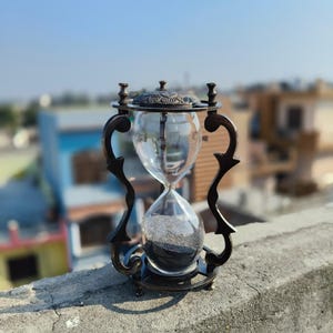 Op de afbeelding: Een antieke zandloper met een donker bronzen frame en heldere glazen bollen. De zandloper bevat zwart zand. Het decoratieve frame heeft gebogen, sierlijke details. De achtergrond toont een heldere blauwe lucht en gebouwen.