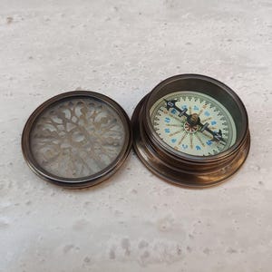 Puede incluir: Una brújula de latón vintage con tapa decorativa. La esfera de la brújula presenta un diseño detallado con marcas direccionales. La tapa está abierta, revelando un intrincado patrón calado. La brújula está sobre una superficie clara y texturizada.