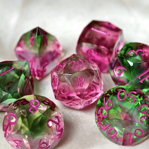May include: A set of polyhedral dice featuring a translucent blend of pink and green colours. Each die has sharp, geometric facets and is engraved with numbers in a bright, bold pink font, including a twenty-sided die showing the number 20.