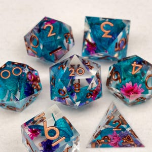 Puede incluir: Un conjunto de siete dados poliédricos transparentes. Cada dado contiene inclusiones florales turquesas y moradas, con números cobrizos. Diseñado para juegos de rol.