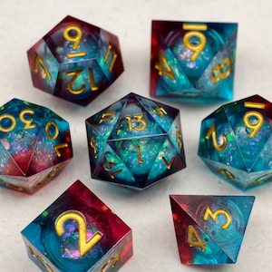 Op de afbeelding: Een set van zeven polyhedrale dobbelstenen met een fantasiethema. De dobbelstenen zijn een mix van blauw, teal en rood met gouden cijfers. De dobbelstenen zijn gemaakt van hars en hebben een glitterachtig, iriserend effect. Perfect voor tabletop gaming.