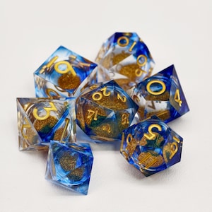 May include: A set of seven polyhedral dice with a clear resin base, infused with blue and gold glitter. The dice feature gold numbering and are designed for tabletop gaming. The set includes a D4, D6, D8, D10, D12, D20, and a percentile die.