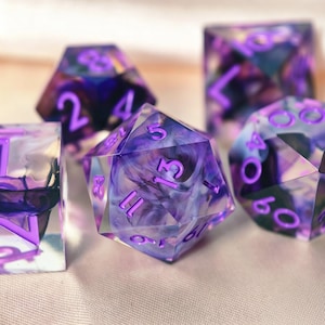 May include: A set of polyhedral dice made from translucent resin with swirling purple and dark blue ink patterns. Each die features sharp edges and bright purple engraved numbers. The collection includes a d20, d12, d8, and d4, all displaying a distinct marbled effect within the clear material.