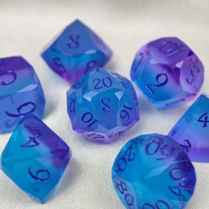 May include: A set of seven polyhedral dice featuring a translucent gradient of blue and purple colours. Each die has sharp, geometric edges and dark blue engraved numbers. The set includes various shapes such as a d20, d12, d10, d8, d6, and d4.