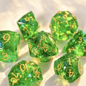 Può includere: Un set di sette dadi poliedrici verde traslucido con numeri color oro. Ogni dado presenta una forma geometrica sfaccettata e contiene piccole inclusioni color oro scintillanti incorporate nel materiale in resina.