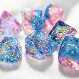 Può includere: Un set di sette dadi poliedrici caratterizzati da una miscela traslucida di tonalità blu e rosa con scaglie di lamina iridescente incastonate. Ogni dado ha bordi affilati e numeri color oro, incluso un dado a venti facce al centro.